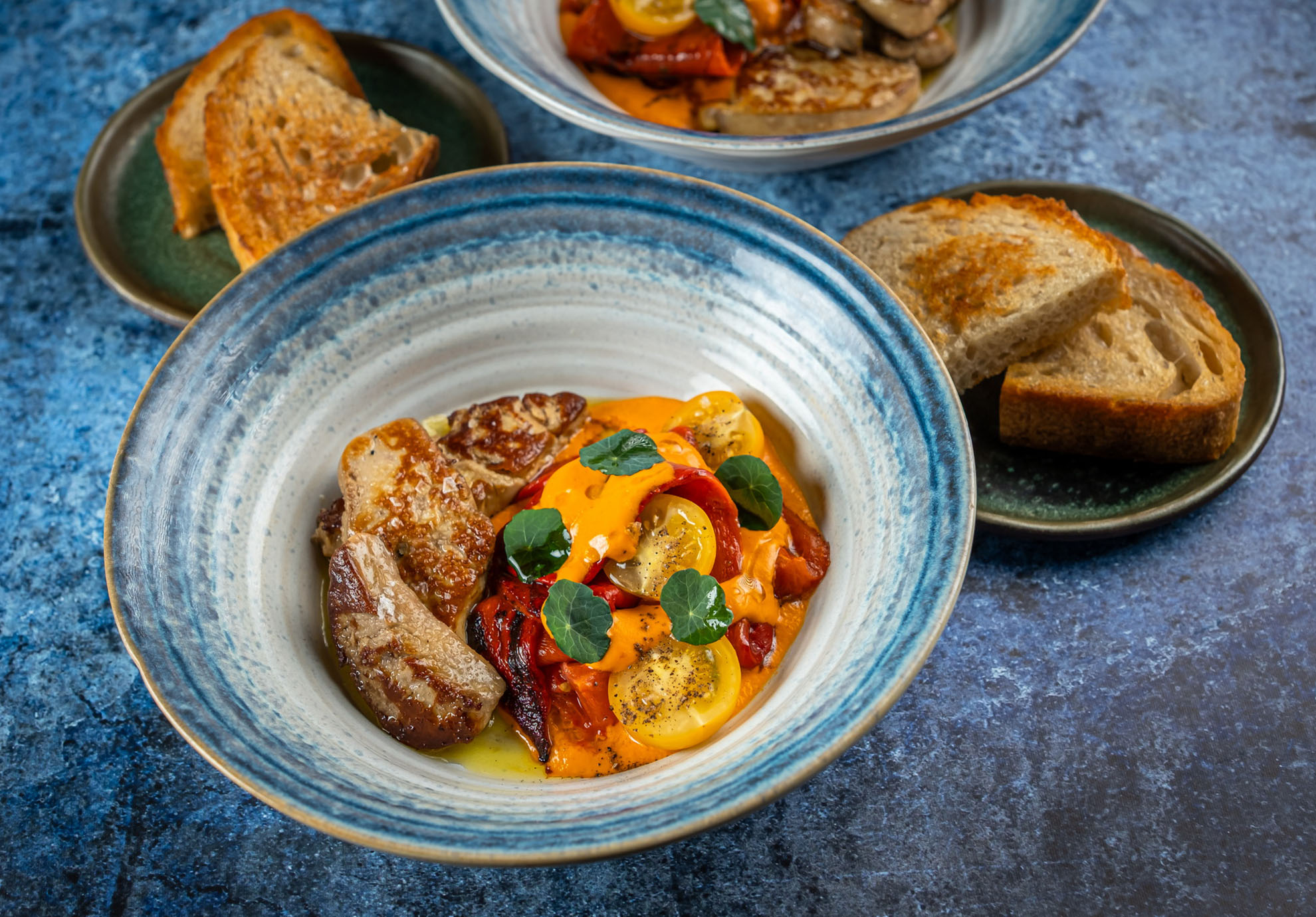 Ein Gericht aus bunten, gebratenen Gemüsesorten, garniert mit Sauce und grünen Kräutern, dazu gegrillte Entenleber auf einem blau-weißen Teller. Rustikale Brotscheiben werden auf einem separaten Teller serviert.
